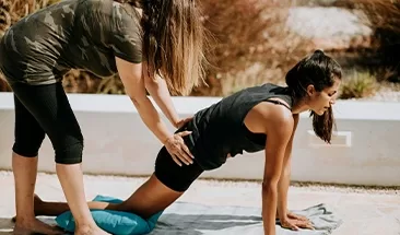 A yoga instructor guiding their client on a yoga pose on a yoga mat in the sunshine.