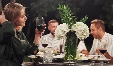 A group of people enjoying a dinner and wine outside sat at a beautifully decorated table with flowers.