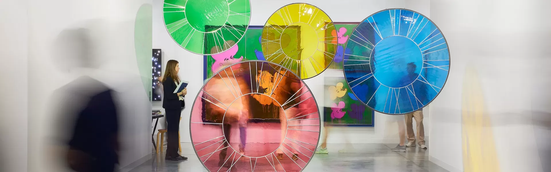 A modern art gallery displays colorful circular stained glass pieces and abstract paintings. Blurred silhouettes of people walk by, enhancing the sense of movement and dynamic energy in the brightly lit space.