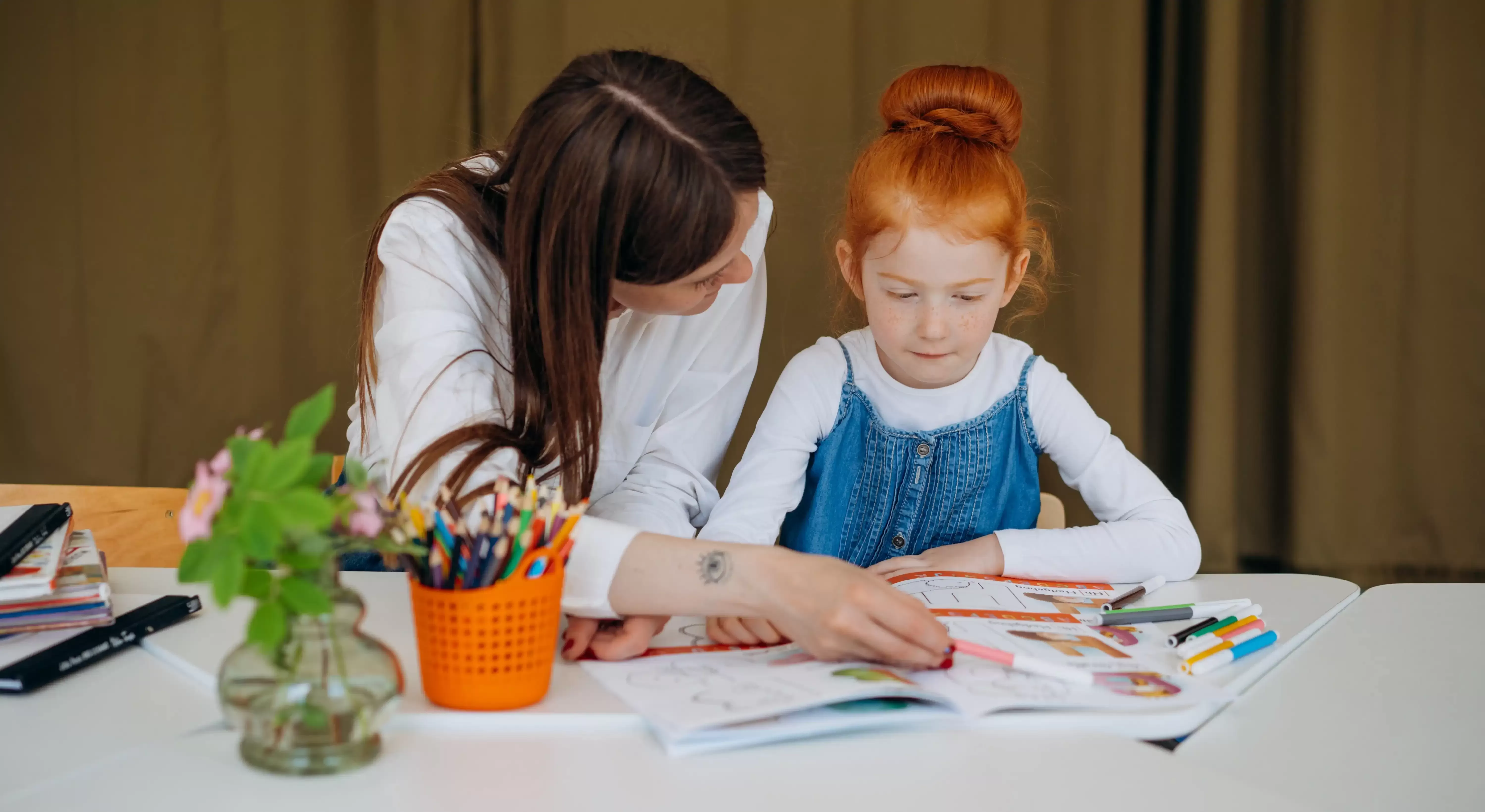 Female teacher sat at table filled with stationary, plat and books helping a young female student