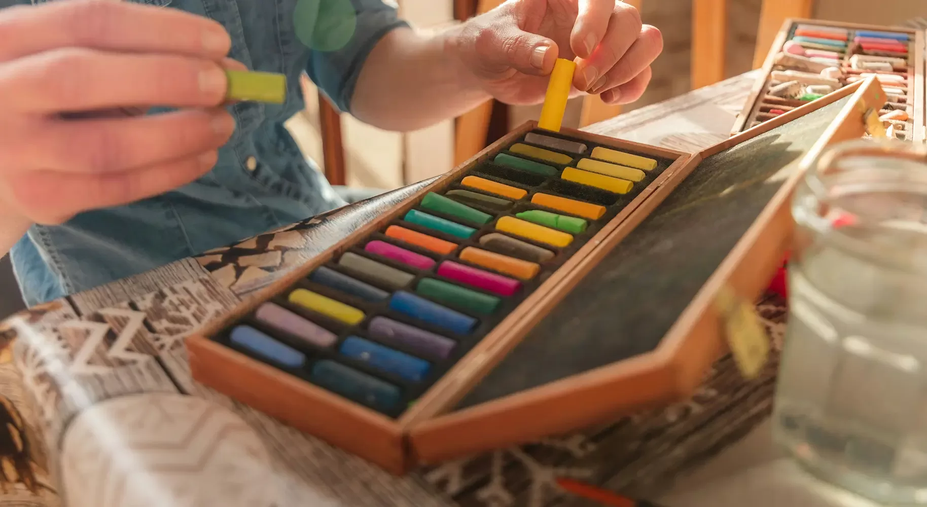 A person selects a yellow pastel stick from a wooden box containing various colorful pastel sticks. The box is on a newspaper-covered table next to a glass jar of water. Sunlight illuminates the scene from the side.