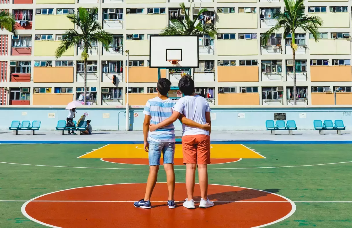 Colourful photo of two boys hugging each other in basketball court looking up at a hoop in front of flat building in the sun