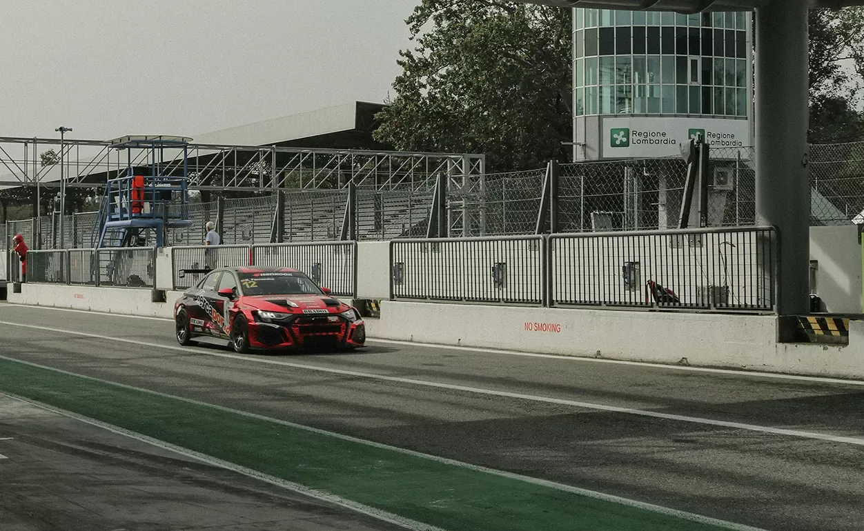 A red and black race car drives down the pit lane of a racetrack, with safety barriers and a glass control tower visible in the background on an overcast day.