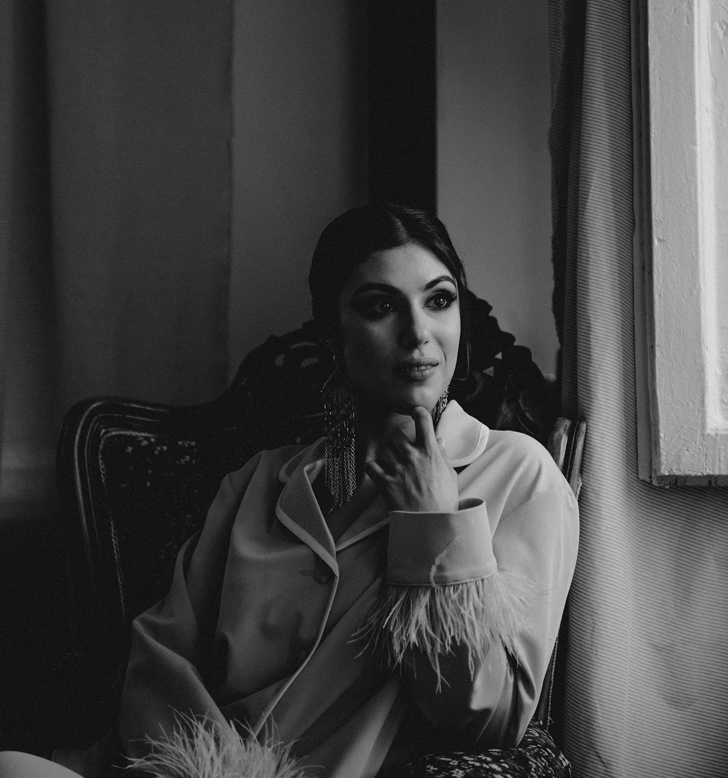 Black and wite photo of a glamorous woman wearing pyjamas with feather sleeves wearing longline statement earrings, sitting on a chair and looking out of a window.