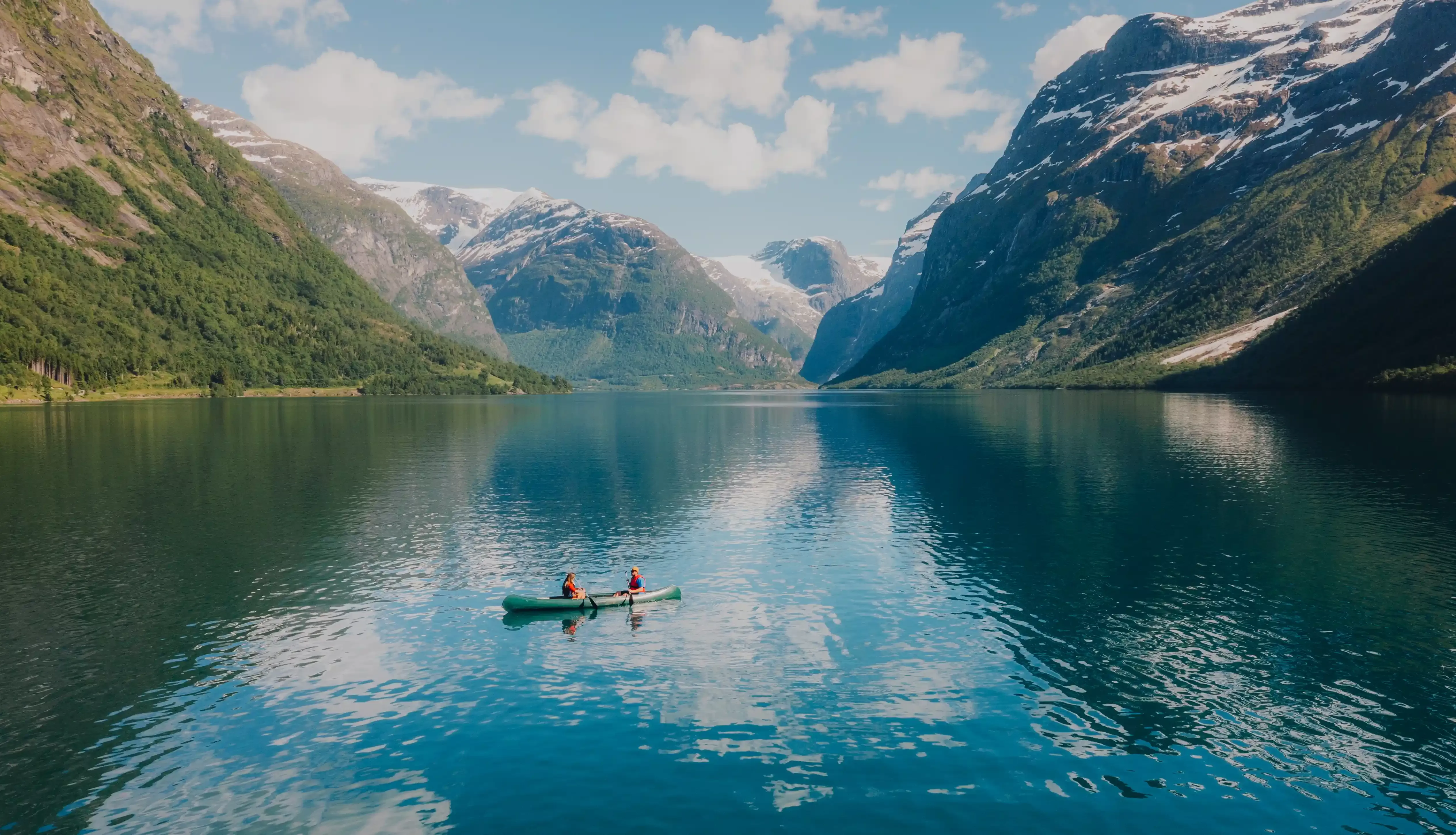 Two people canoe on a calm, clear lake surrounded by majestic mountains with snow-capped peaks under a partly cloudy sky. The reflections of the mountains and sky are visible on the waters surface.