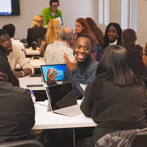 A group of people seated at tables in a classroom setting, engaging in lively discussion. Some have laptops in front of them. A person in a light green shirt stands at the front, possibly leading the session. Everyone appears focused and engaged at a Quintessentially Foundation event.