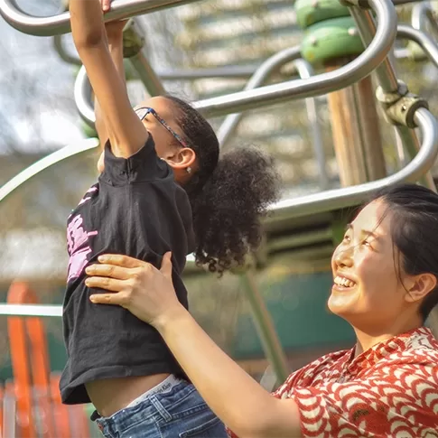 A female child swinging on monkey pars at the park being held up by a lady in a red dress, smiling - by Quintessentially Foundation.