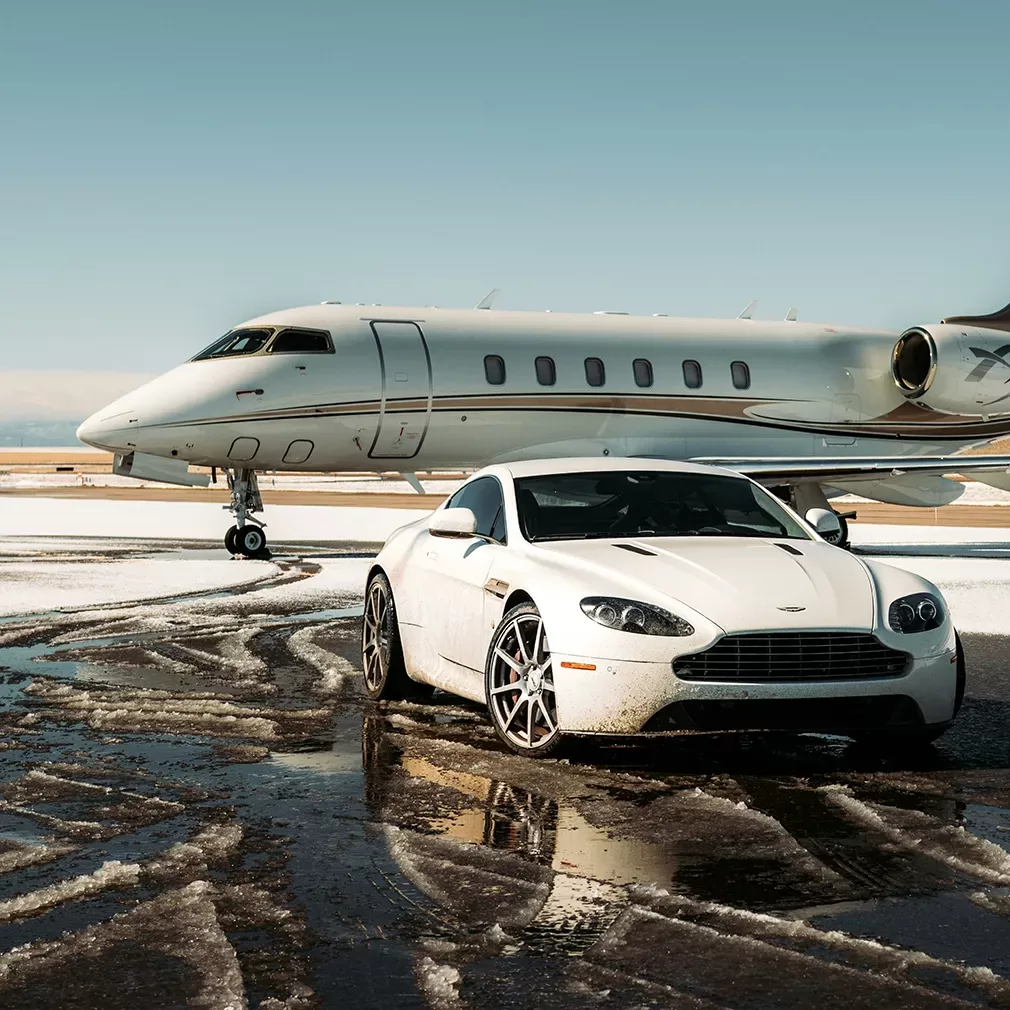 A white luxury sports car is parked on a wet, snow-dusted runway in front of a sleek private jet under a clear sky.