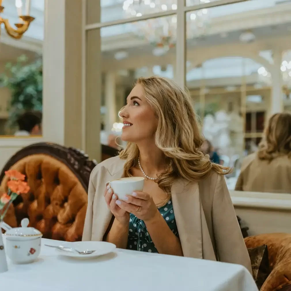 A woman with wavy blonde hair sits at an elegant café table, holding a teacup and smiling as she looks upward. She wears a beige coat over a blue dress and is surrounded by plush seating and ornate mirrors.