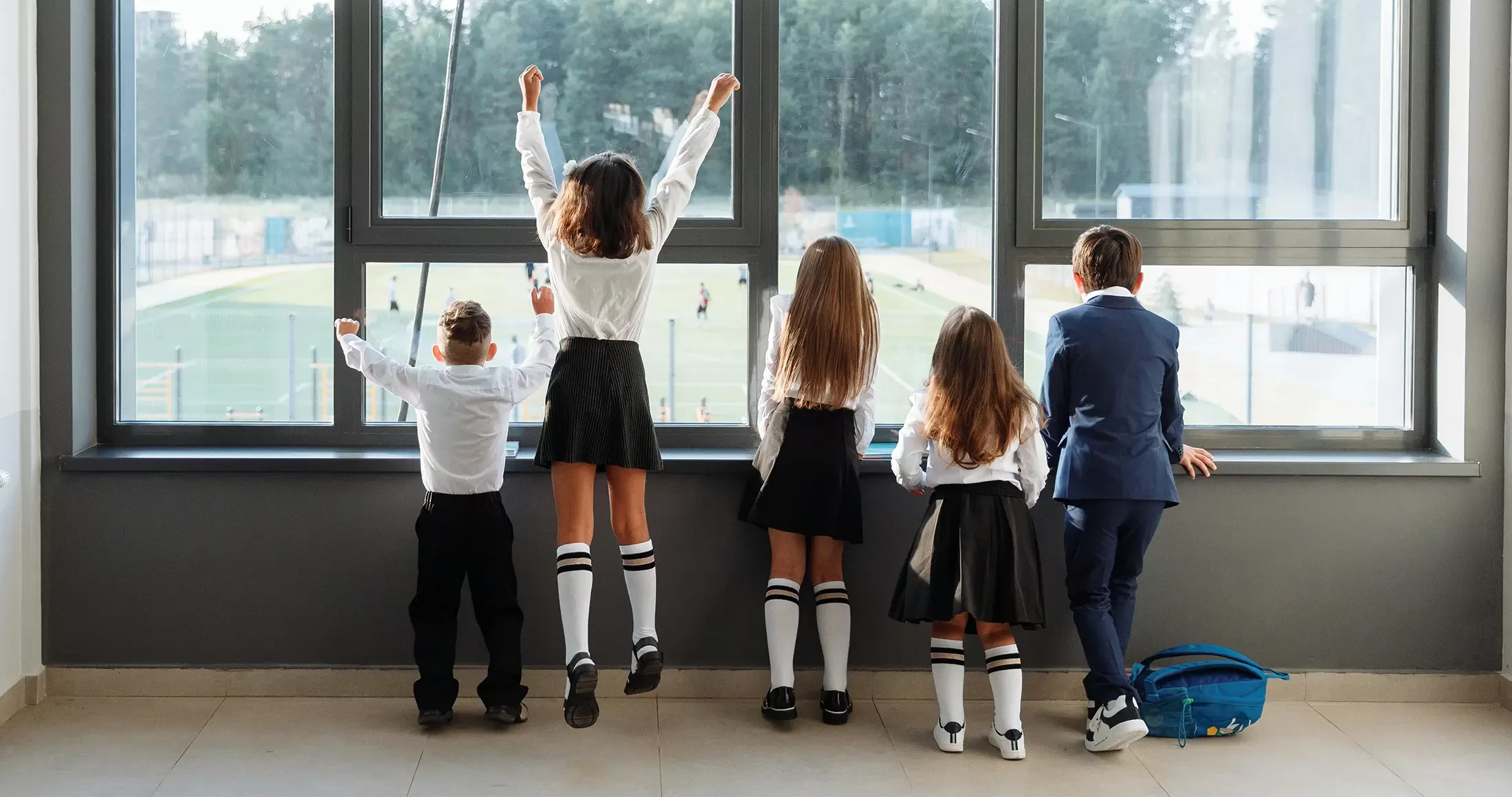 Five young children in school uniforms stand by a large window, looking outside. Three girls in black skirts, white shirts, and knee-high white socks stand alongside two boys, one wearing a suit and the other in a white shirt and black pants. The scene outside shows a green field with trees in the background, and some buildings are visible in the distance.