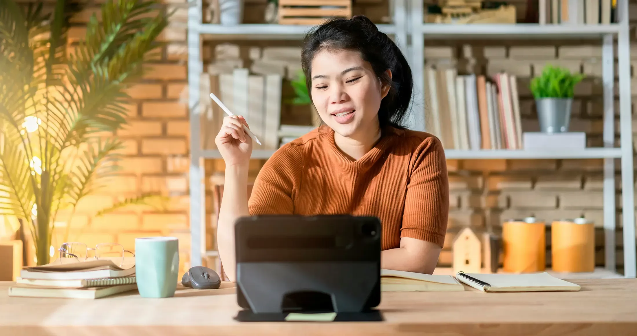 A woman sitting at a wooden desk in a well-lit home office, smiling as she looks at a tablet screen. The desk is neatly organized with books, a cup of coffee, and a mouse. The background features shelves filled with books and plants, creating a warm and inviting workspace. The woman is casually dressed in an orange top, holding a stylus pen, ready to work or study.