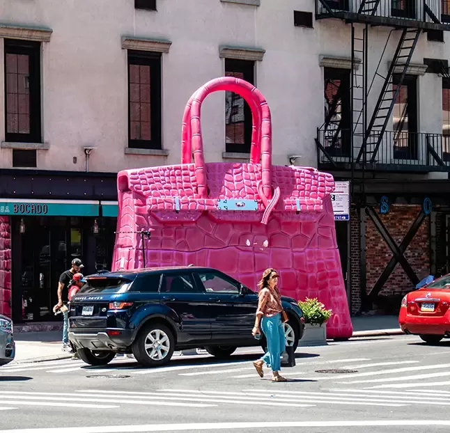 A woman walks past a giant pink handbag sculpture on a city street. Cars are parked nearby, and the sculpture is positioned in front of a building with multiple windows and a fire escape. The scene is bright and sunny.