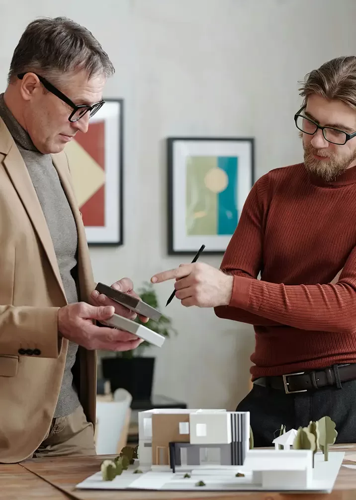 Two men in an office discuss over a table with an architectural model on it. One wears a brown jacket and holds a book, while the other in a red sweater gestures with a pen. Framed abstract art is visible on the wall behind them.