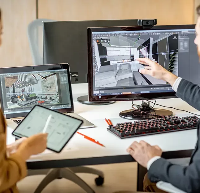 Two people work at a desk with multiple computer screens displaying architectural design software. One points at a monitor showing 3D building models, while the other holds a tablet. A keyboard and a notebook are also on the desk.