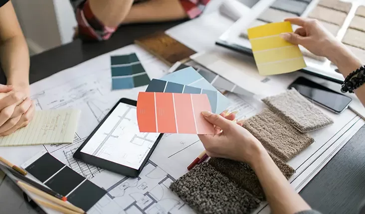 Three people working on a design project, holding color swatches over architectural plans. A tablet and samples of fabric and carpet are on the table, suggesting interior design or renovation work.