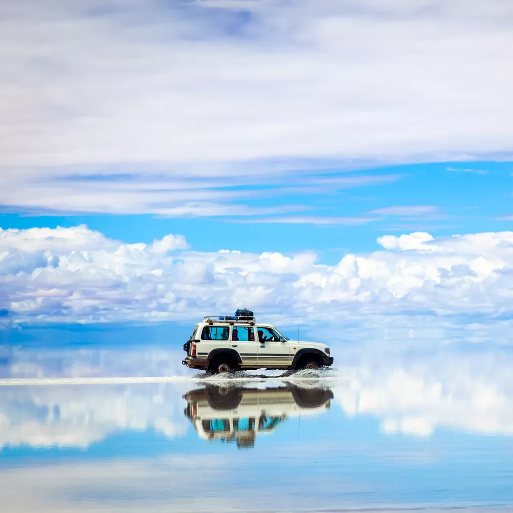 A white SUV drives across a reflective, water-covered salt flat under a blue sky with scattered clouds, creating a mirror-like effect on the ground.
