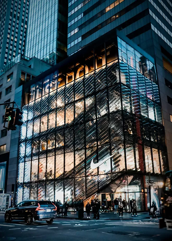 A bustling city street at night features a brightly lit, multi-story Nike store with a glass facade. People walk on the sidewalk, and cars drive by under the reflection of surrounding skyscrapers.