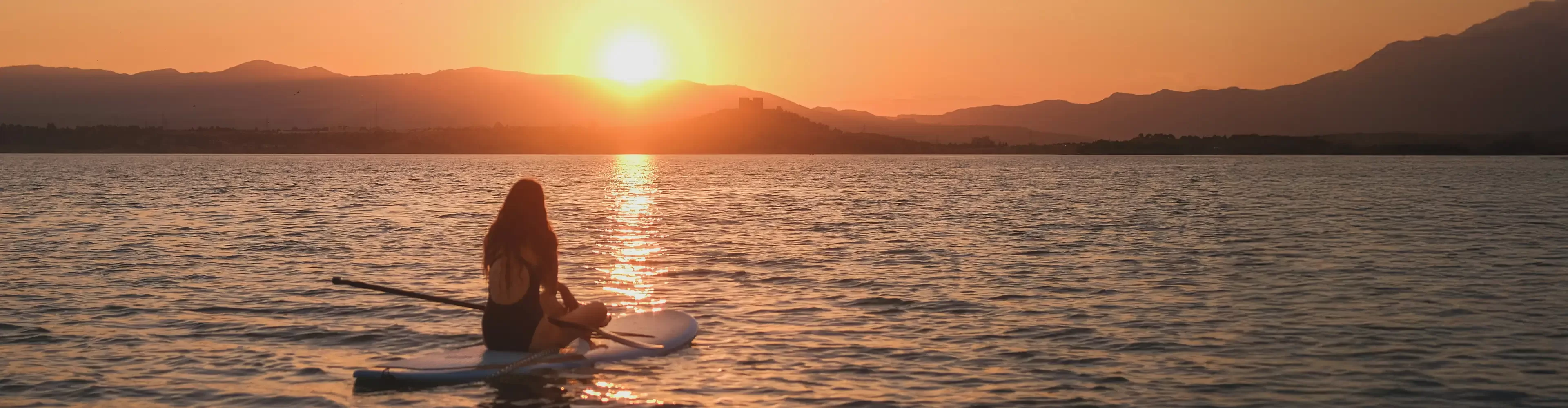 A person sitting on a paddleboard on calm water at sunset, with the sun low over distant mountains, casting a golden reflection across the lake.