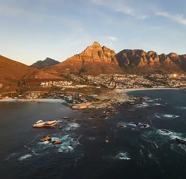 A scenic view of a coastal city at sunset with rugged mountains in the background. The coastline features rocky shores and a calm sea, while the city nestles between the mountains and the water. The sky is clear with a warm, golden hue.