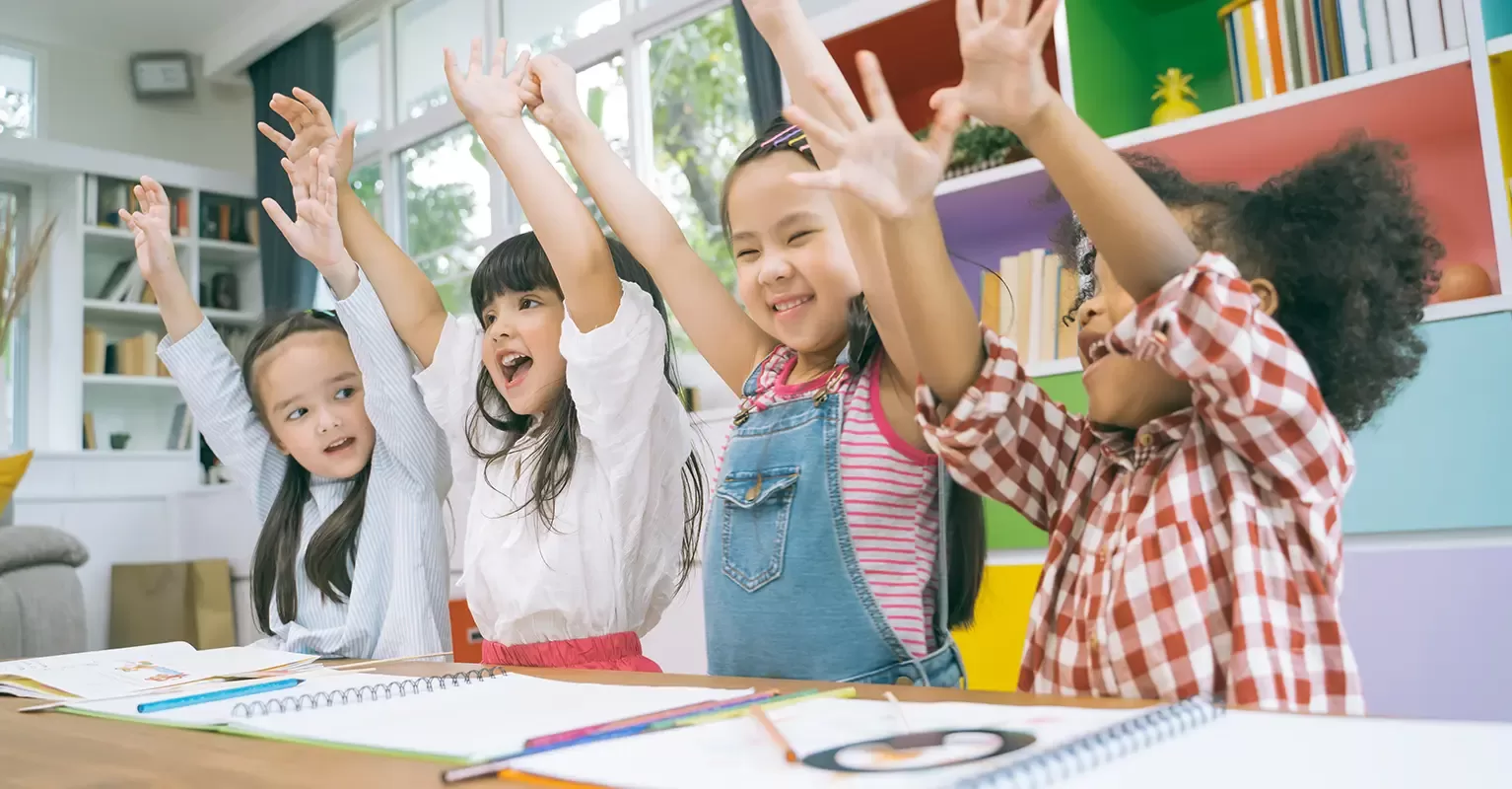 Four young children sit at a table in a brightly colored classroom, smiling and raising their arms enthusiastically. Drawing supplies and open notebooks are on the table in front of them.