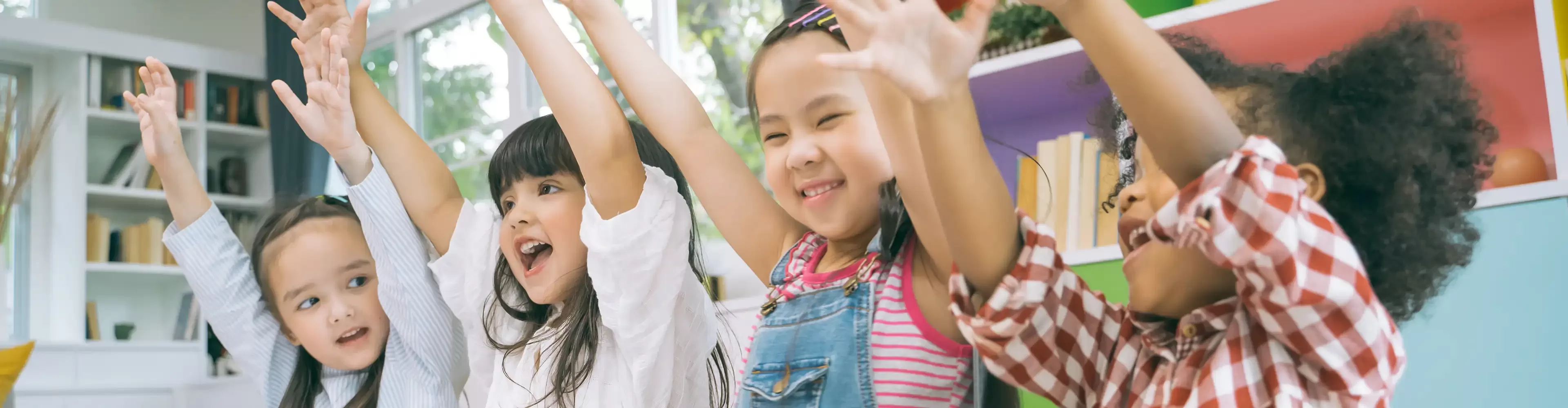 Four young children smile and raise their arms in excitement while sitting indoors, surrounded by bright shelves and books, creating a cheerful and playful atmosphere.