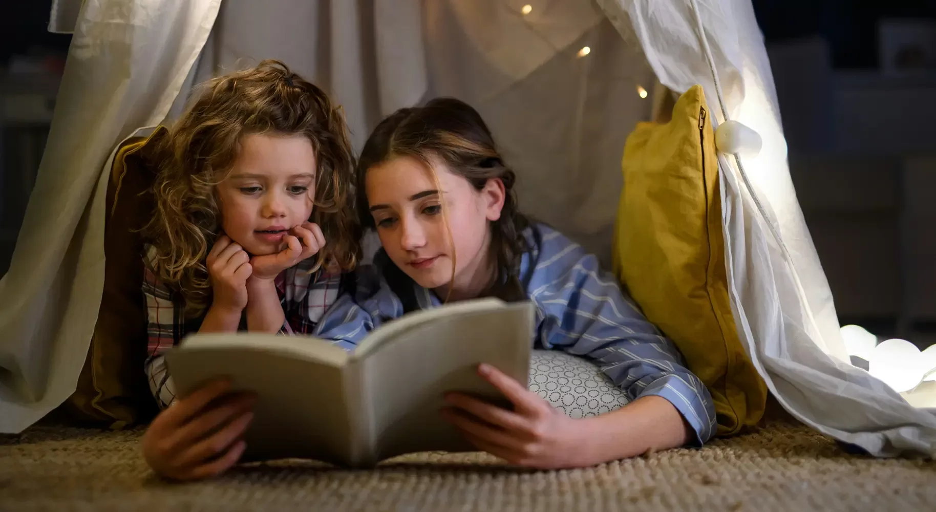 Two children are lying on their stomachs inside a cozy indoor tent, reading a book together. They are propped up on pillows, with soft lighting creating a warm atmosphere. String lights adorn the inside of the tent.