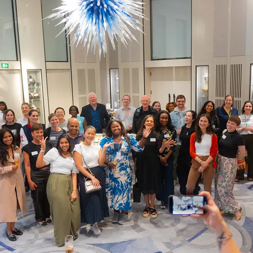 A diverse group of people pose for a photo in a large, well-lit room with a striking blue and white chandelier overhead. They are smiling, some holding drinks, while one person in the foreground takes a picture with a smartphone by Quintessentially Foundation.