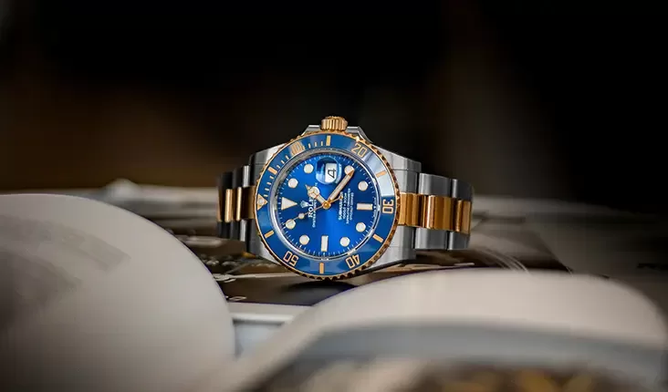 A luxury wristwatch with a blue dial and gold accents is placed on an open magazine. The watch features a rotating bezel with gold and blue colors, and a stainless steel and gold bracelet. The background is softly blurred.