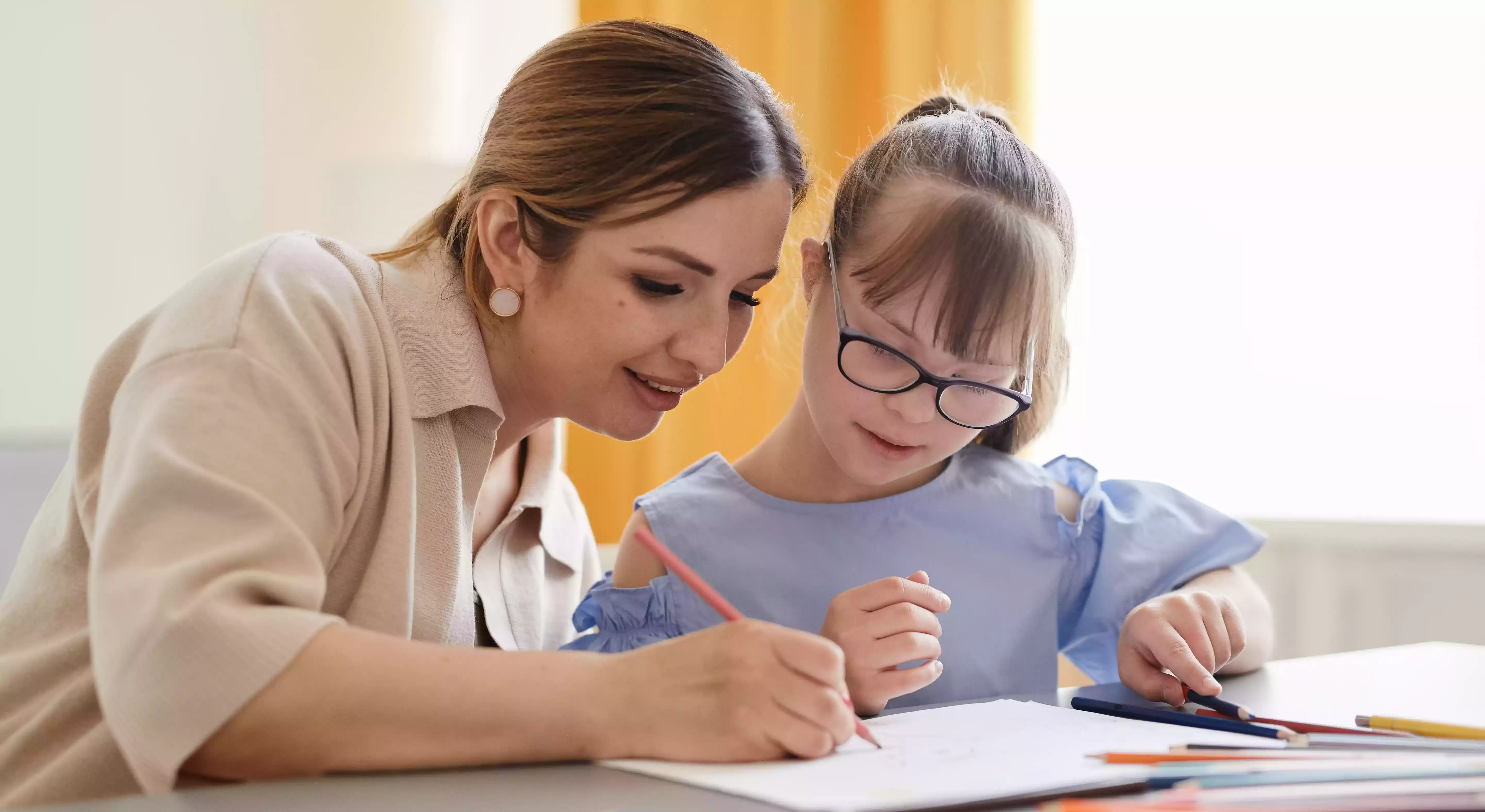 Girl with special needs writing in a book with support of female teacher