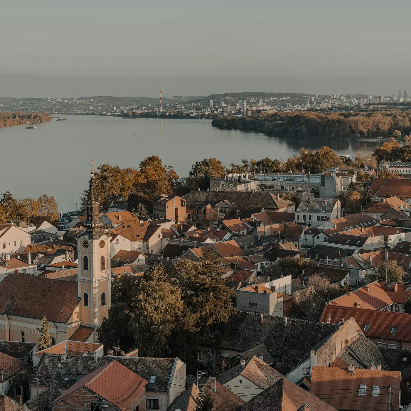 Aerial view of a town with red-roofed buildings and a tall church tower in the foreground. The town is bordered by a wide river on the left, with a tree-lined shore and a cityscape visible in the distance under a clear sky.