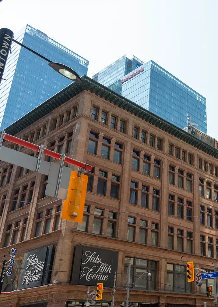 Street view of a historic building with Saks Fifth Avenue signs, set against a backdrop of modern glass skyscrapers. Traffic lights and street signs are visible in the urban scene.