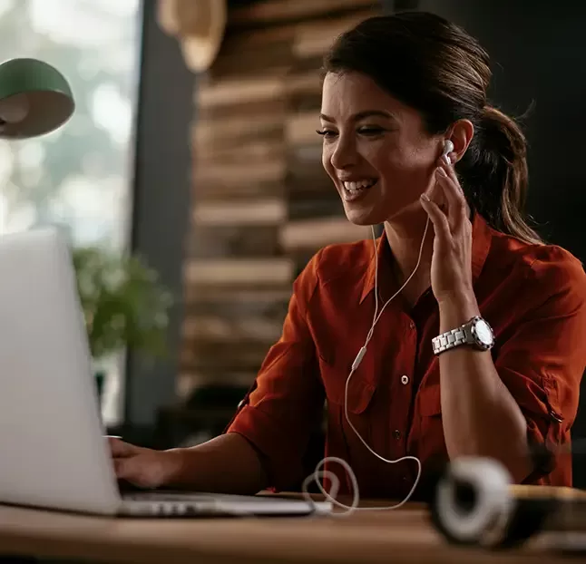 A woman wearing a red shirt smiles while using a laptop. She has earphones in and is seated at a desk with some blurred background elements, suggesting a cozy and well-lit environment.