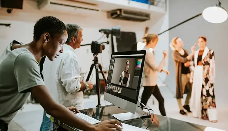 A person works on a computer in a photography studio, editing an image on screen. In the background, a photographer and an assistant prepare two models for a photo shoot, with lights and cameras set up around them.