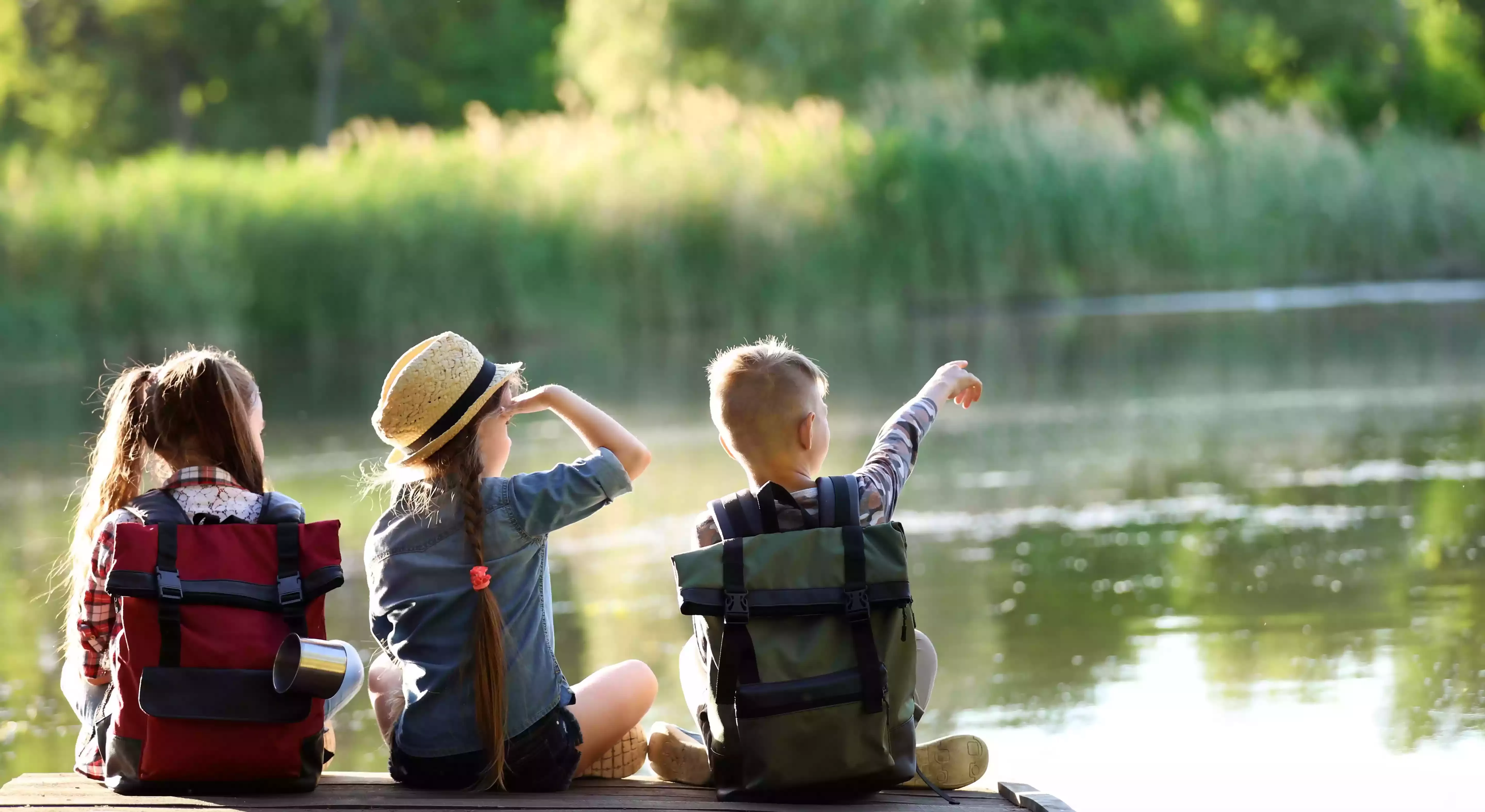 Three kids sat with rucksacks in school uniform on the side of a lake in the sun looking up at the sky