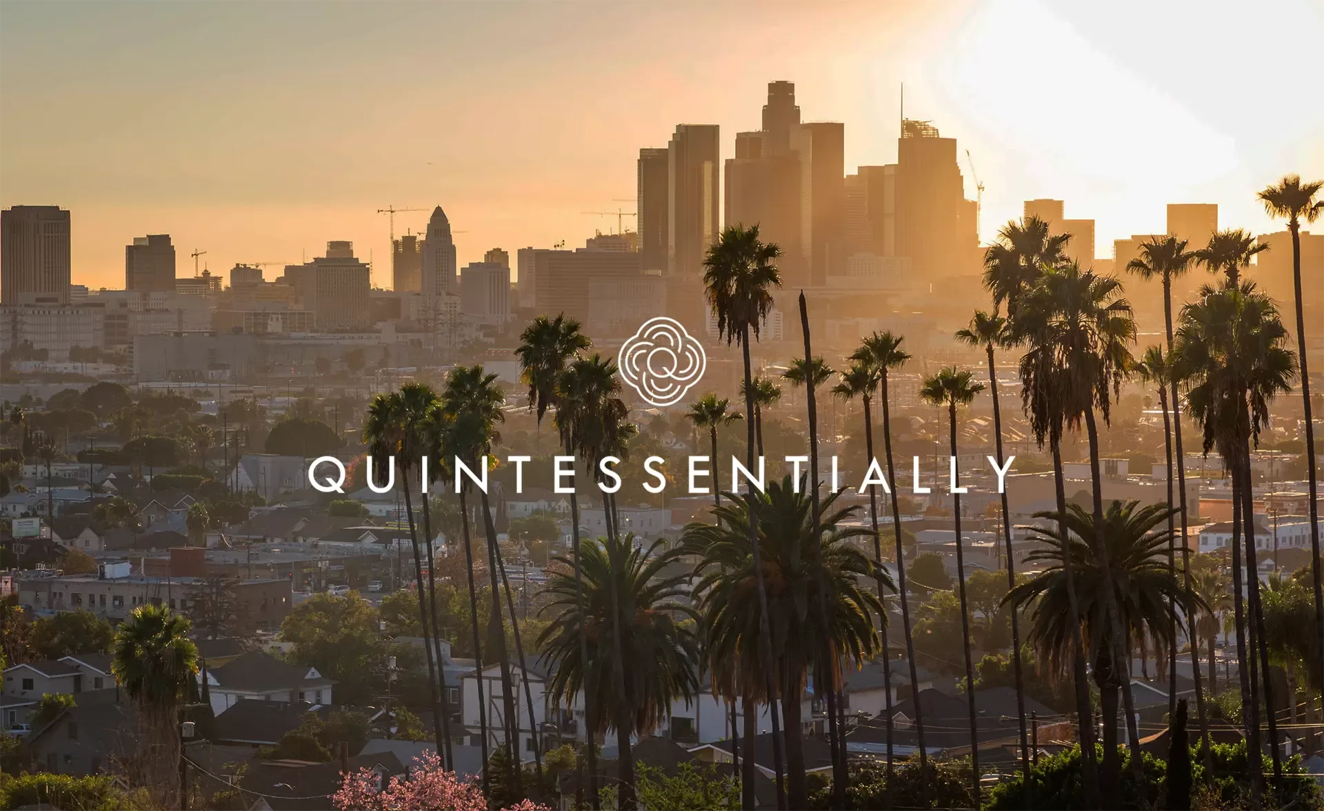 A sunset view of downtown Los Angeles with palm trees in the foreground. The city skyline is silhouetted against the golden sky. Text and a logo in the center read QUINTESSENTIALLY.