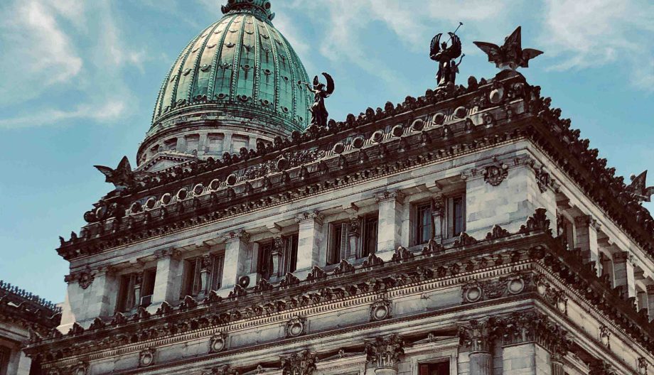 The image shows a close-up of a grand neoclassical building with a prominent green dome. The facade features intricate sculptures and decorative elements, including statues of figures on the rooftop. The sky is clear and blue in the background.