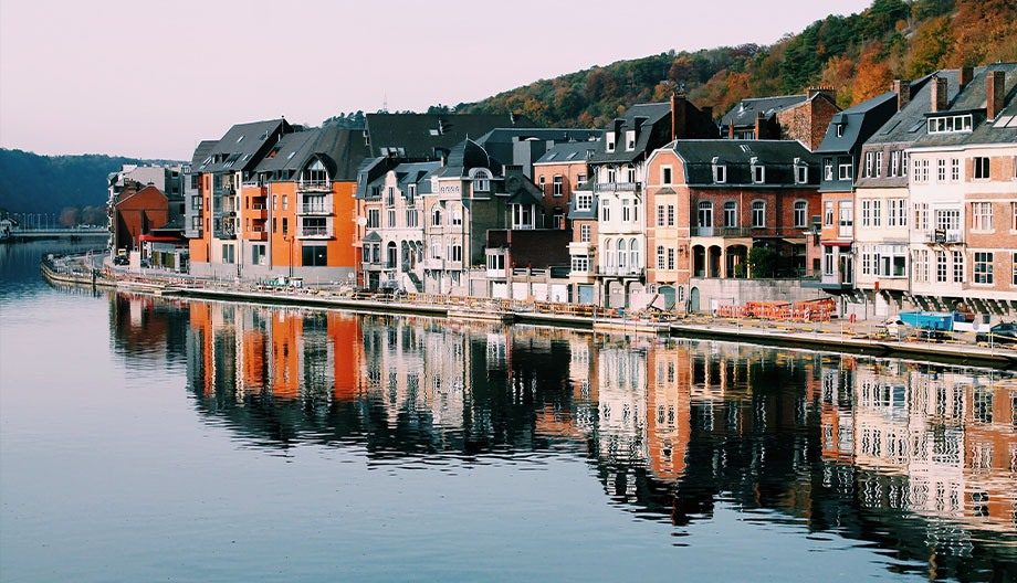 Quaint European riverside town with charming brick and stone buildings lining the waterway. Their reflections create a mirror image on the still water. In the background, trees with autumn foliage cover the hillside.
