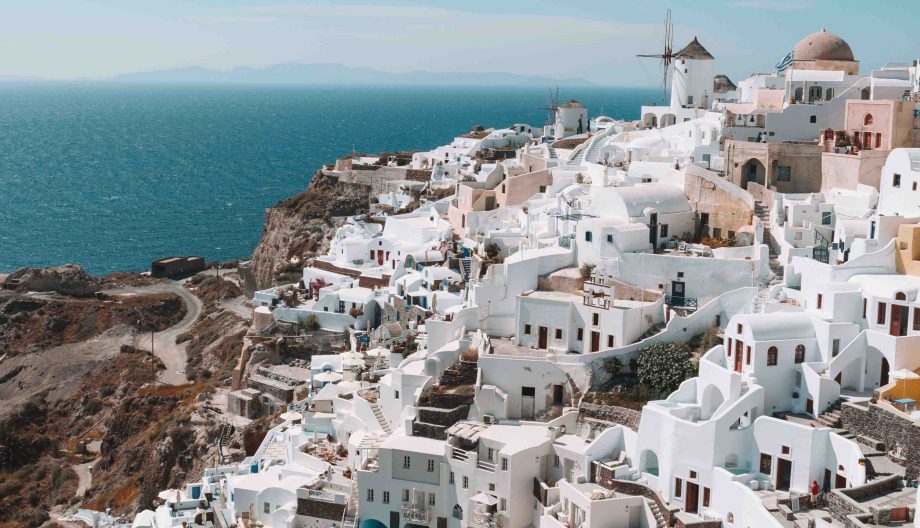 A seaside Greek village with whitewashed buildings and blue domes cascades down a hillside toward the sea. A traditional windmill is visible, and the sea extends to the horizon under a clear blue sky.