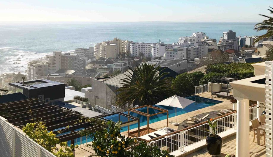 Scenic view of a coastal city with modern buildings and a vast ocean in the background. A pool and lounge area with an umbrella and palm tree occupy the foreground, surrounded by a railing.