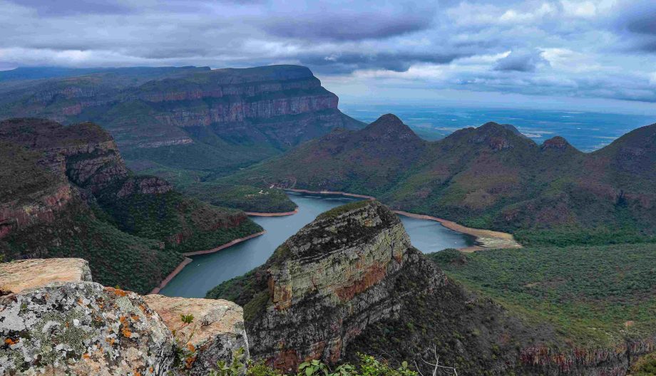 Scenic view of the Blyde River Canyon in South Africa, featuring lush green cliffs and mountains surrounding a winding river under a cloudy sky.
