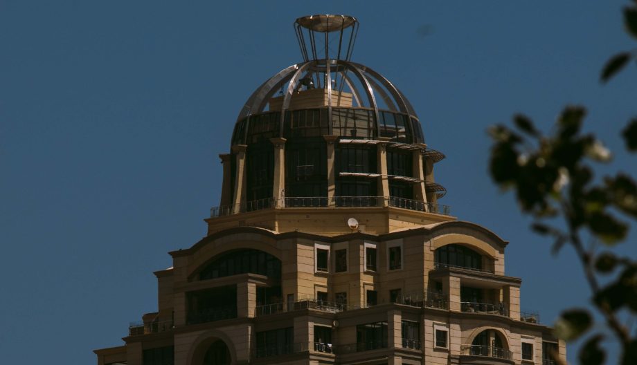 The image shows the top portion of a beige building with a distinctive dome structure against a clear blue sky. The dome features a metal framework, and parts of the building have balconies. Some blurred tree leaves are visible in the foreground.