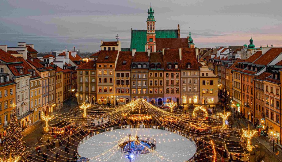A picturesque view of a festive town square at dusk, featuring a large ice rink surrounded by illuminated string lights. Historic buildings with red roofs frame the scene, with a prominent church in the background.
