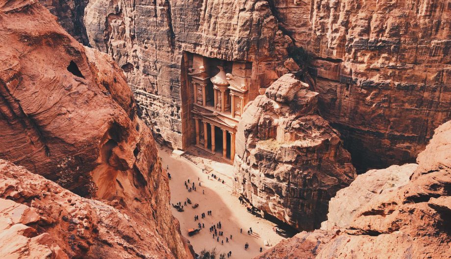 Aerial view of Petras iconic Treasury, an ancient structure carved into red sandstone cliffs. Tourists are gathered below, surrounded by towering rocky walls under a warm, clear sky.