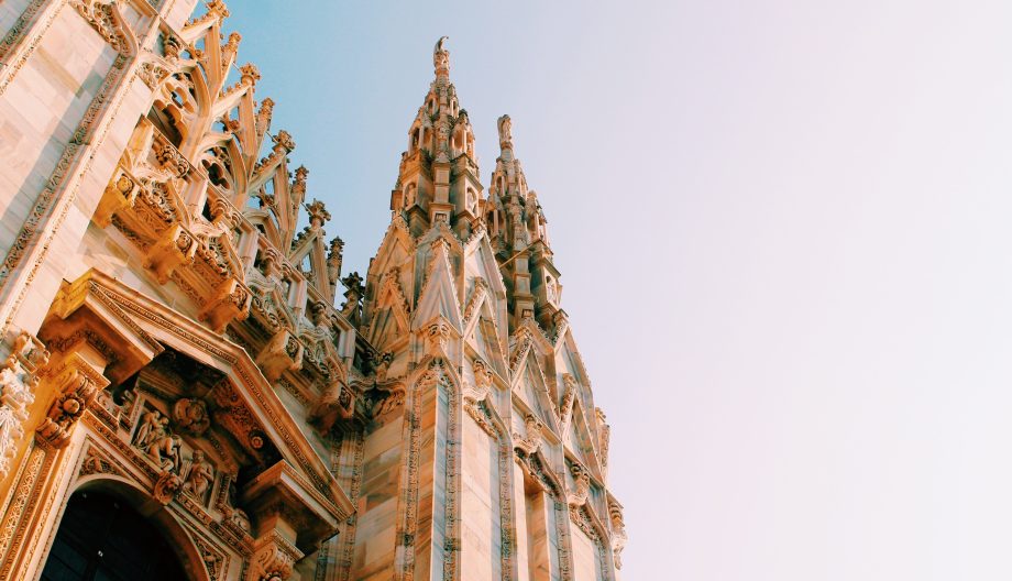 Gothic cathedral spires reach up into a clear blue sky, showcasing intricate stone carvings and architectural details. The sunlight softly illuminates the ornate features of the building.