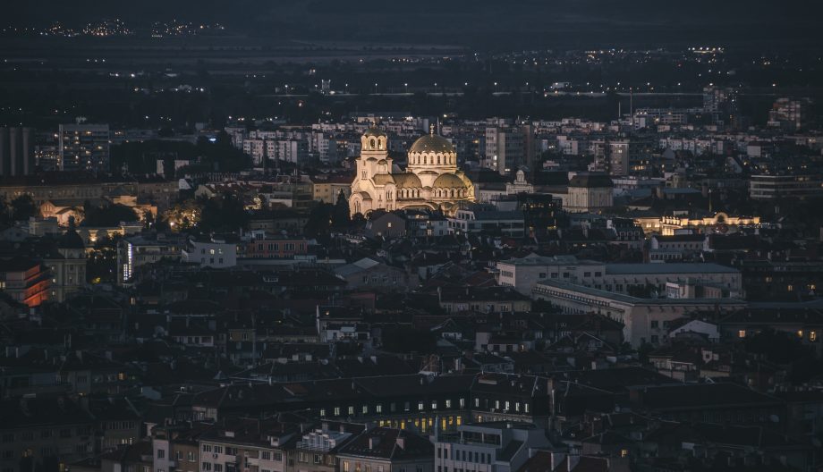 Aerial view of a city at night, featuring a prominently illuminated cathedral with twin domes. The surrounding area is filled with various buildings, all under a dark, expansive sky.