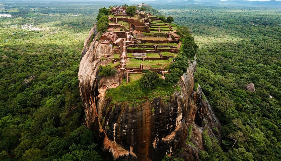 Aerial view of Sigiriya Rock in Sri Lanka. The ancient rock fortress stands tall with visible ruins on top, surrounded by lush green forest. The landscape stretches out into the distance under a clear sky.