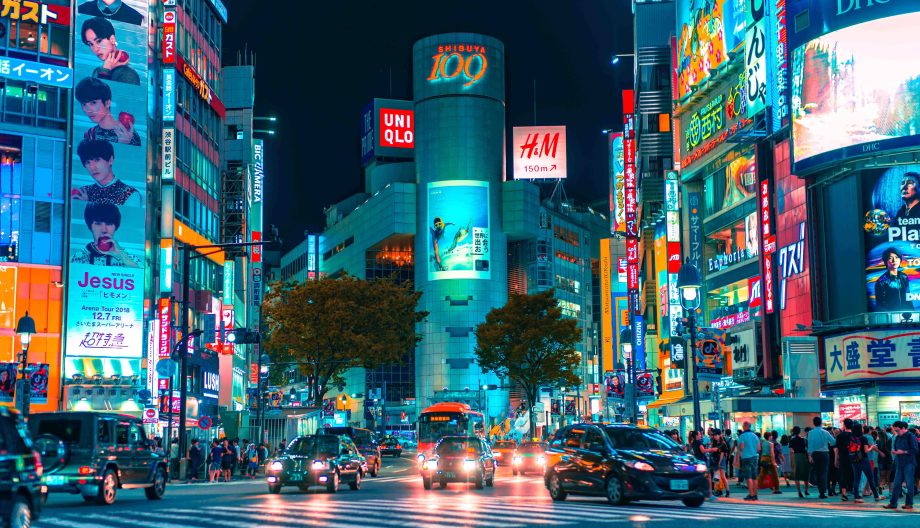 A bustling city street at night, filled with bright neon signs and billboards. People walk on sidewalks, and cars line up at a crosswalk. The scene is vibrant and lively, with tall buildings adorned with various advertisements.