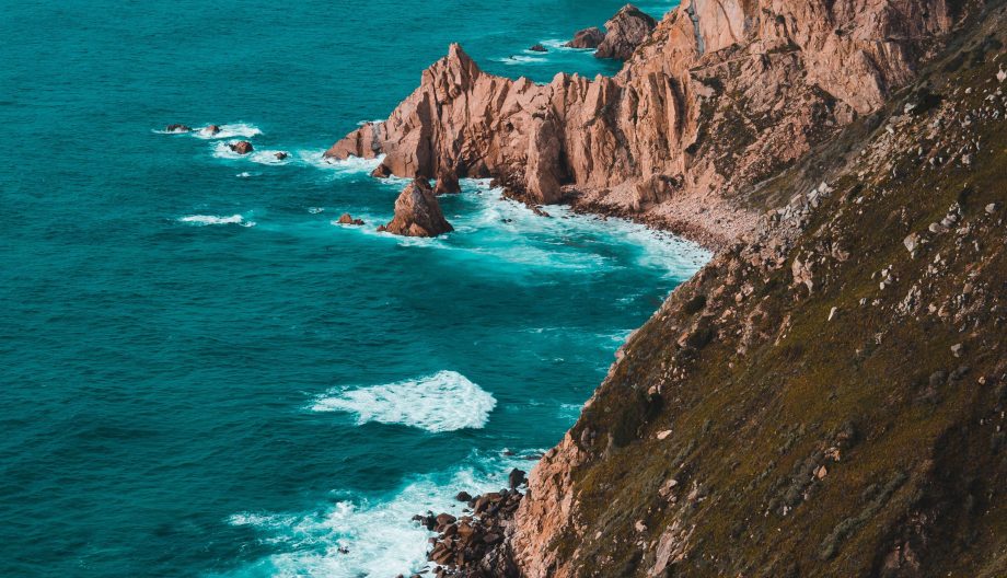 A scenic view of rugged cliffs meeting the ocean, with waves crashing against the rocks. The coastline extends into the distance under a partly cloudy sky, creating a dramatic and natural landscape.