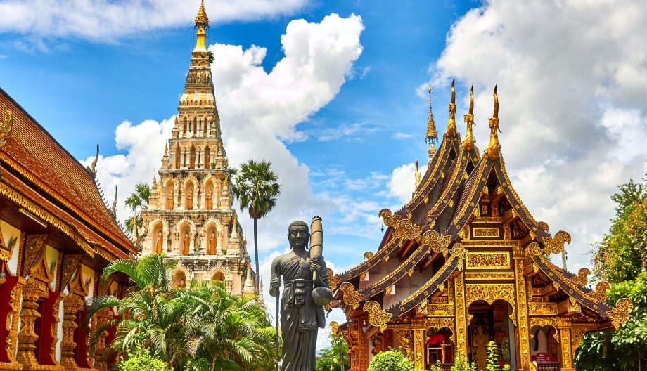A vibrant image of a traditional Thai temple with intricately designed roofs and gold detailing. A statue stands nearby, surrounded by lush greenery. In the background, a tall stupa reaches into a bright blue sky with scattered clouds.