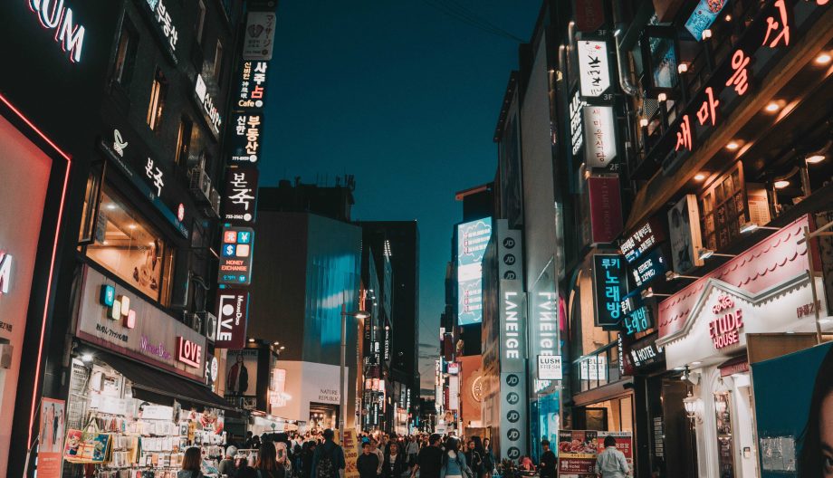 A bustling city street at night, illuminated by vibrant neon signs in various languages. People walk along the sidewalk, and shops line both sides of the street, contributing to the lively urban atmosphere.